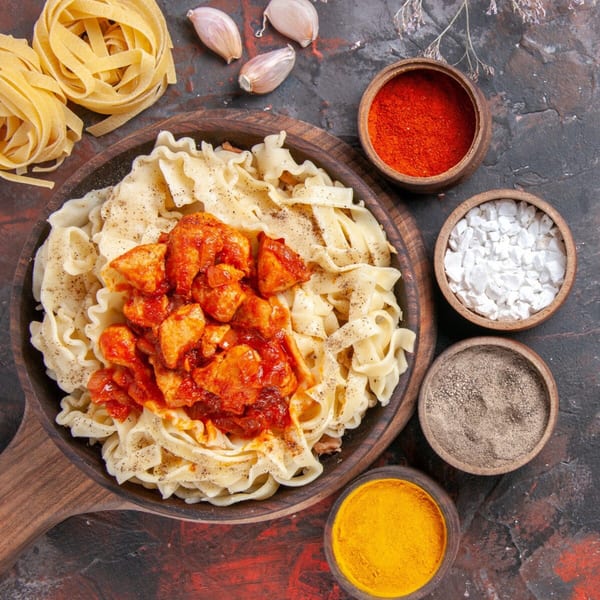 Nutinis' pasta with chicken and red sauce, surrounded by spices, garlic, and uncooked pasta.